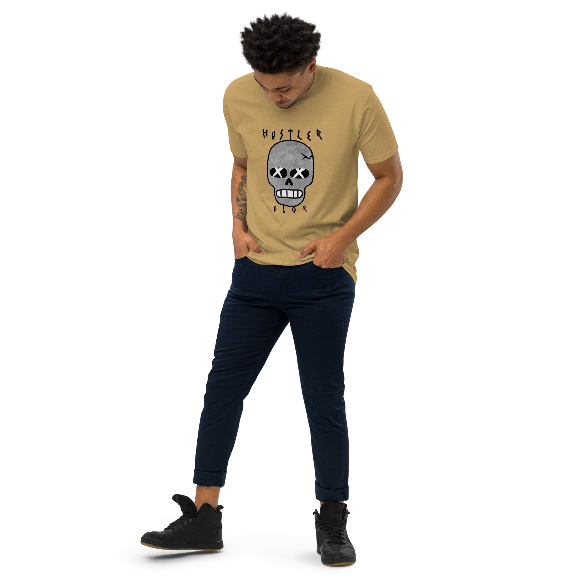 A man in a tan t-shirt with a skull graphic and navy pants stands in front of a white background, looking down.